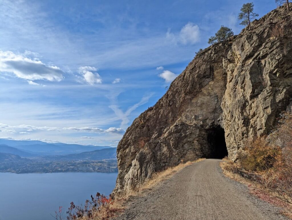 Gravel trail leading through blasted rock tunnel on right, with views of Okanagan Lake below