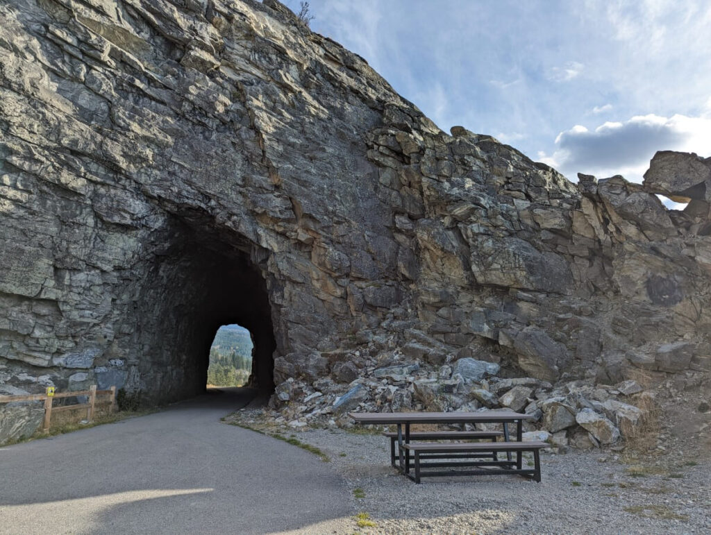 Picnic bench on gravel space next to paved path leading through lasted rock tunnel in Naramata