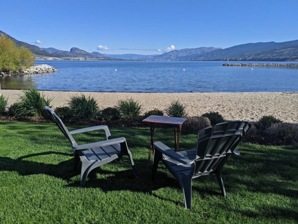 Two chairs and a table perch on grass behind golden sand beach at Marina Way in Penticton. The lake is calm in the background and is backdropped by forested mountains