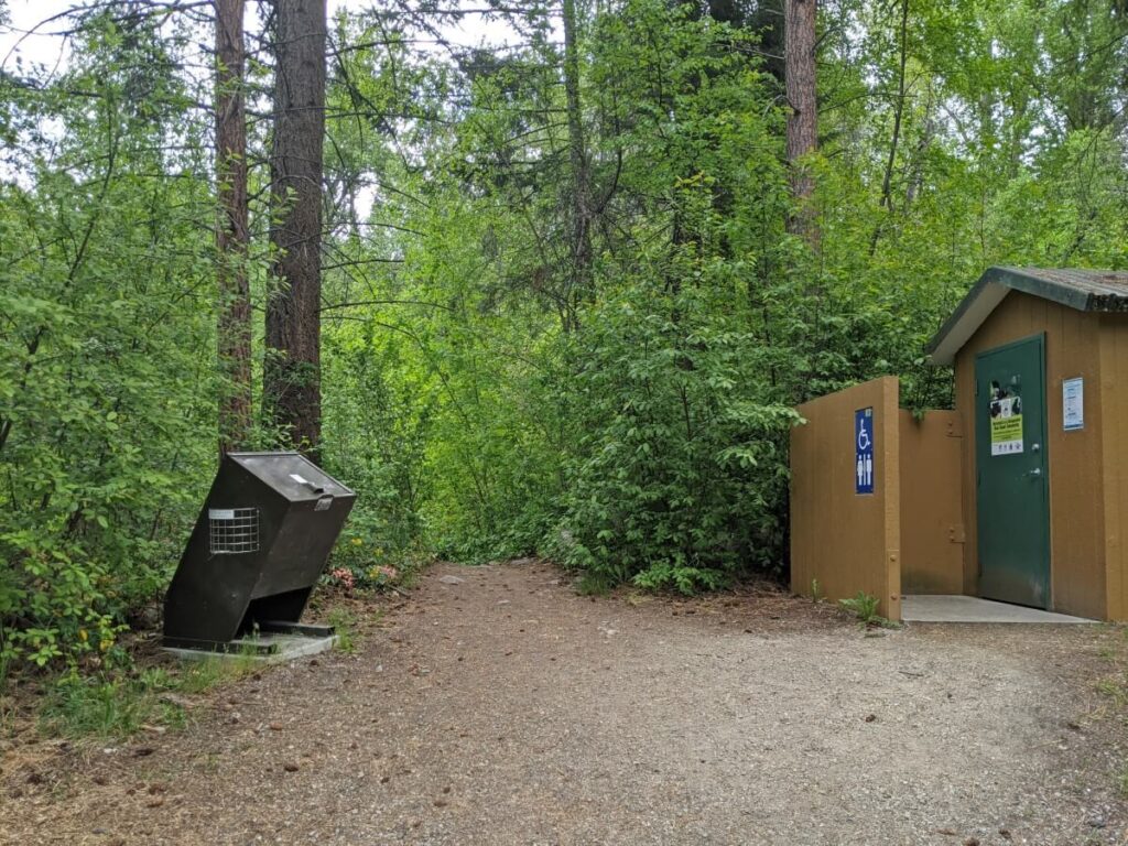 Metal trash bin next to brown outhouse building in Naramata Falls Creek parking area, with backdrop of forest