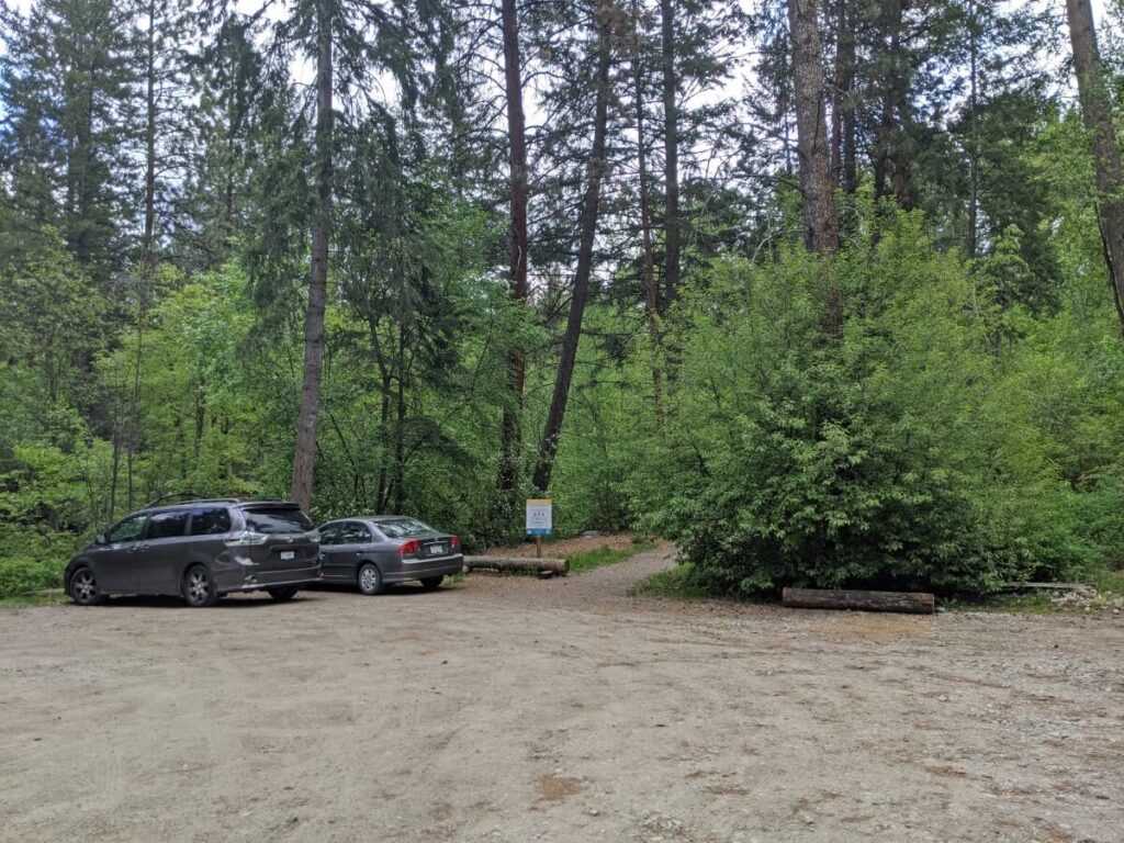 Naramata Creek Falls parking area, with two parked vehicles to the left. The dirt parking lot is surrounded by forest