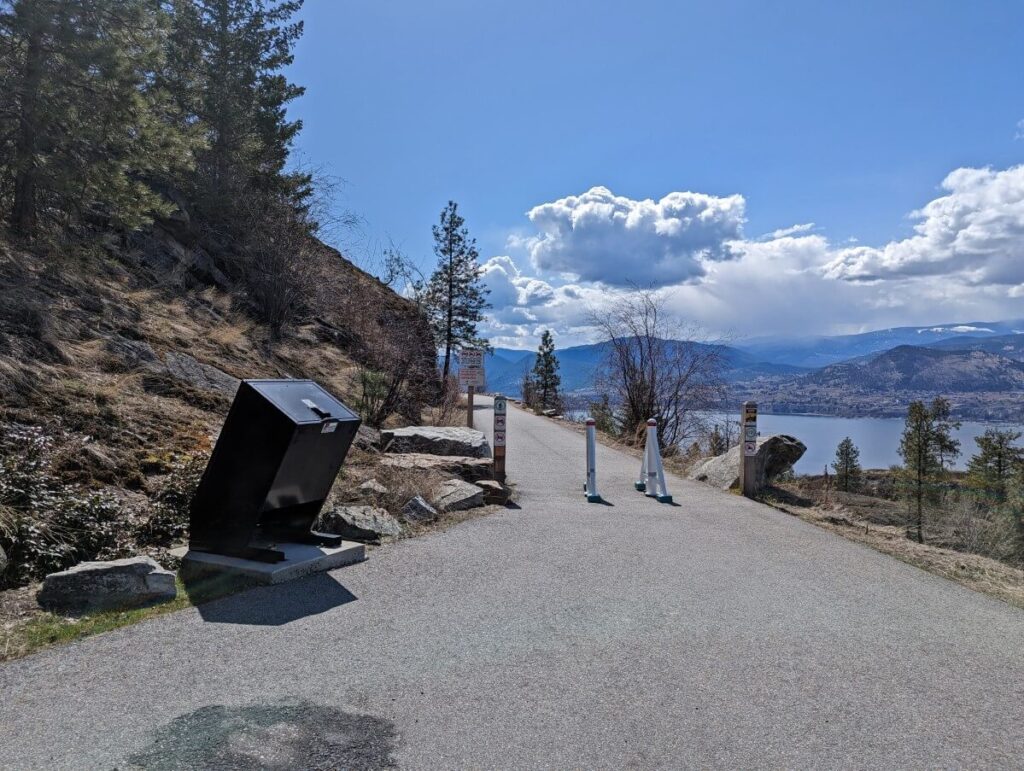 Paved trail area near Little Tunnel with trash bin on left and barrier on right. Okanagan Lake is visible in the background
