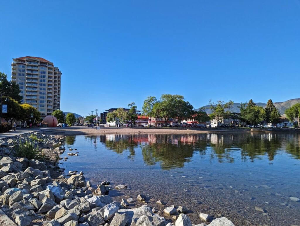 Looking across rock beach next to very calm lake, with golden sand beach visible across water. A large tower building is on the left, with a giant peach sculpture below it. Trees and buildings line the rest of the shore