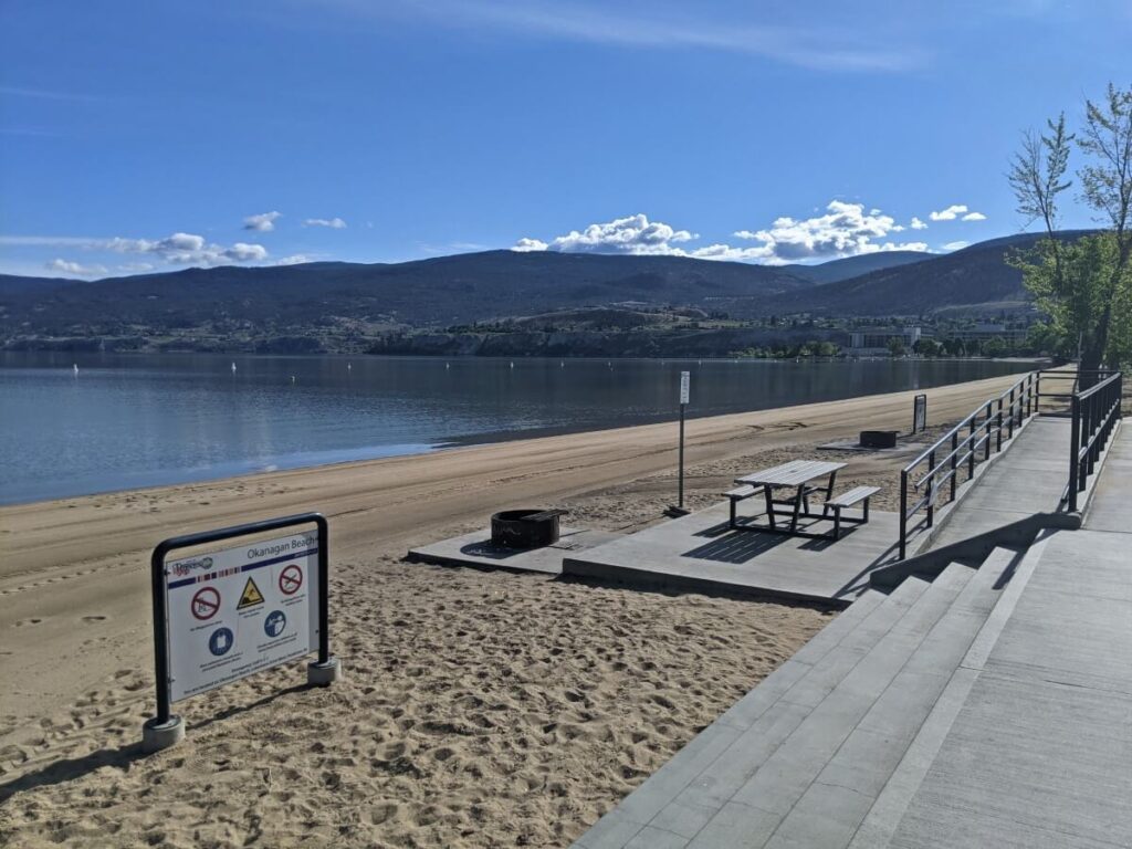 Looking down from paved promenade to paved picnic table on Okanagan Lake beach with steps and ramp access 