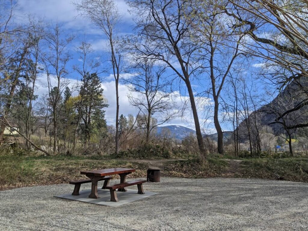 Flat gravel campsite at campground, with wooden picnic table and metal firepit, surrounded by trees and forested hills behind