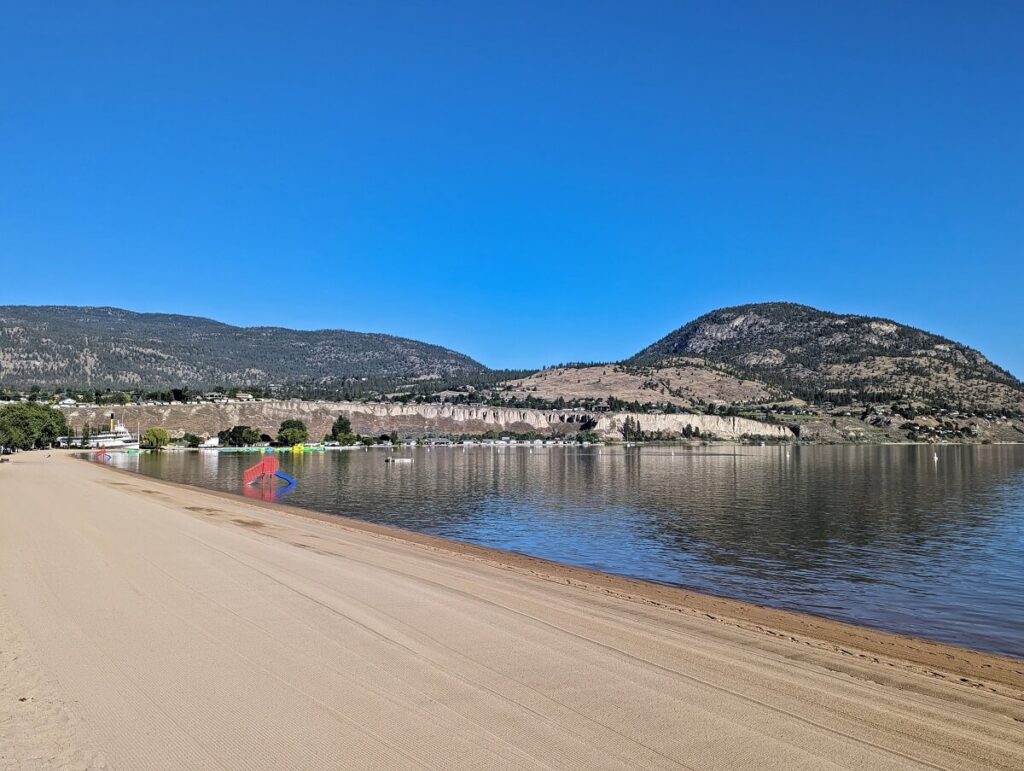 Side view looking down pristine Okanagan Beach in Penticton, with manicured stretch of golden sand. A red and blue slide is visible close to shore. Forested hills in the background. The lake is very calm