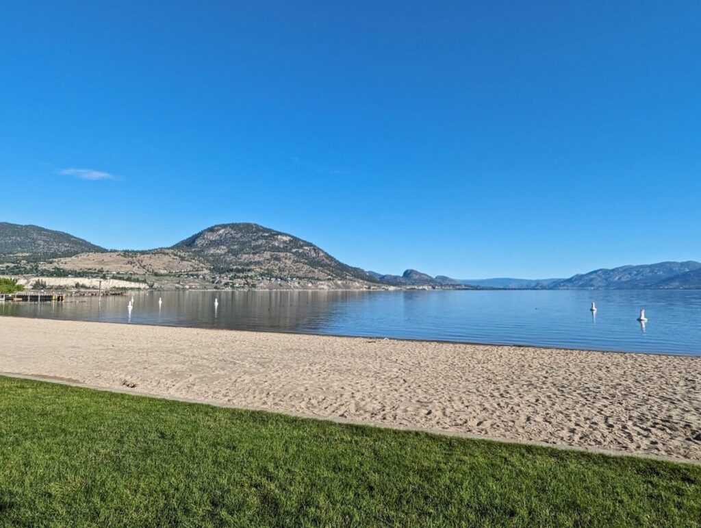 Looking across grass bordered golden sand beach at Okanagan Lake Park, with calm lake in background. A large mountain rises out of the water to the left