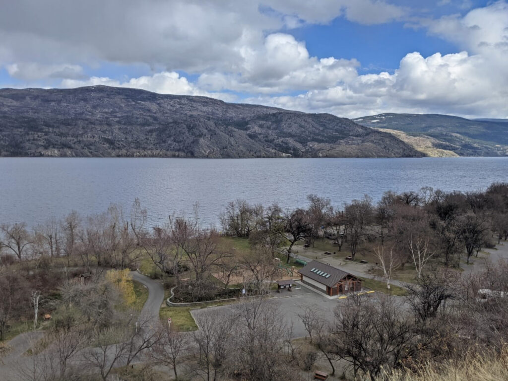 Looking down on Okanagan Lake Provincial Park South Campground, with large wooden shower building visible in front of lawn and campsite area. Okanagan Lake is visible in the background, with rugged park behind