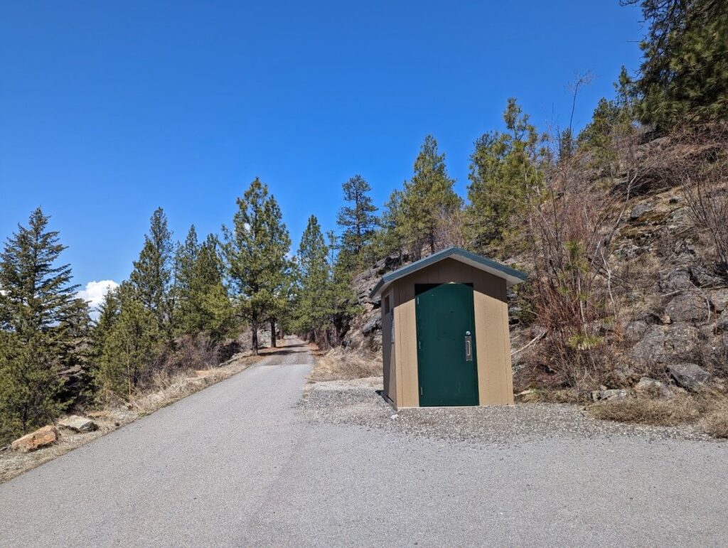 Wooden green and brown outhouse building on gravel area to right of paved KVR Trail