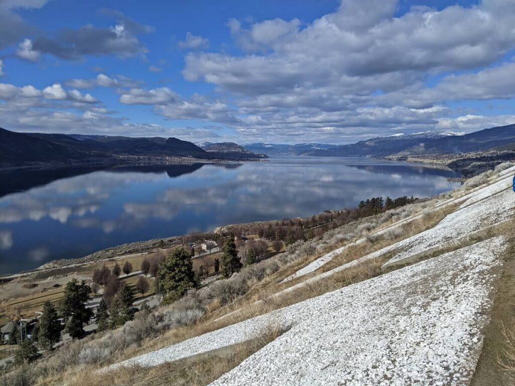 Looking down on huge white Penticton letters on the side of Munson Mountain, with reflective Okanagan Lake visible below. There are forested hills with some snow in the background