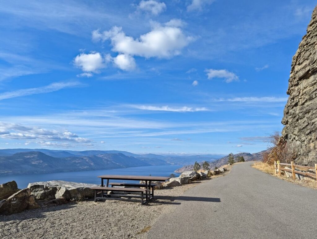 Picnic table on gravel area next to paved KVR Trail at Little Tunnel viewpoint in Naramata. Okanagan Lake is visible in the background