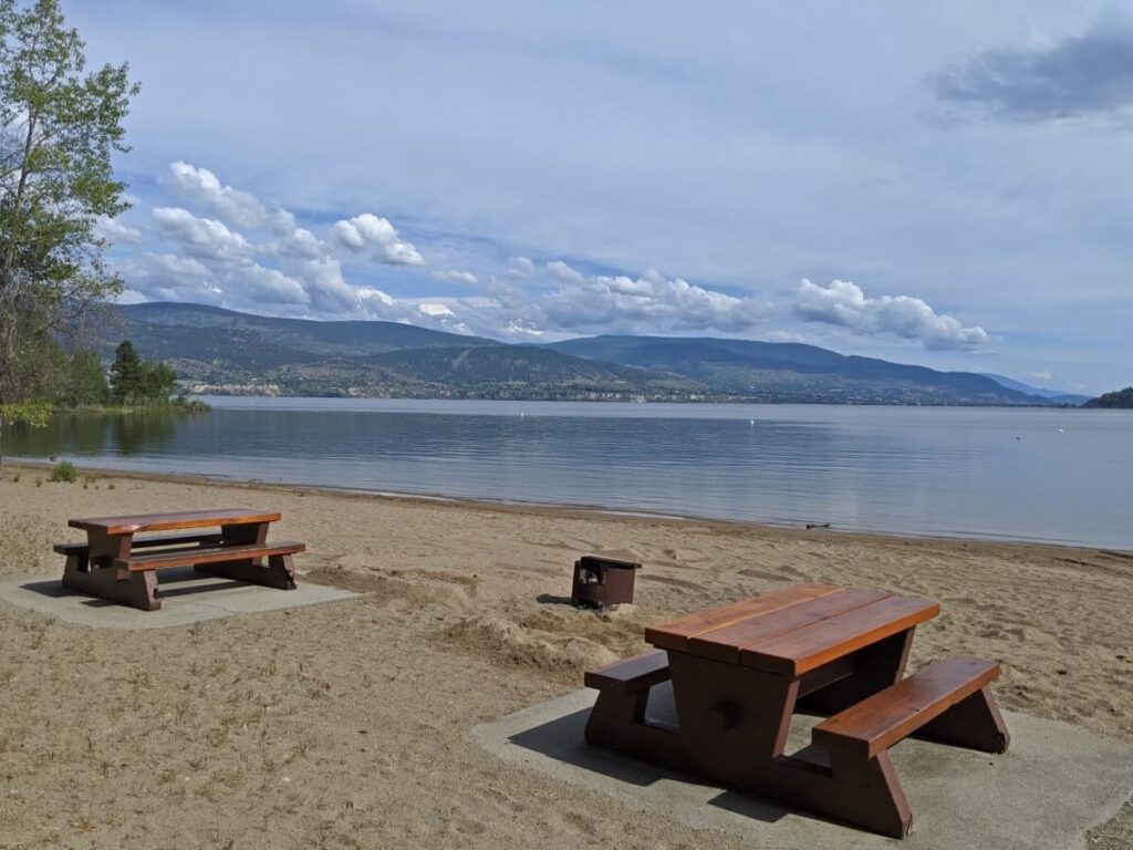 Looking across golden sand beach, with two wooden picnic tables near camera. There is a metal fire pit inbetween the two picnic tables. The lake is very calm in the background and forested hills can be seen