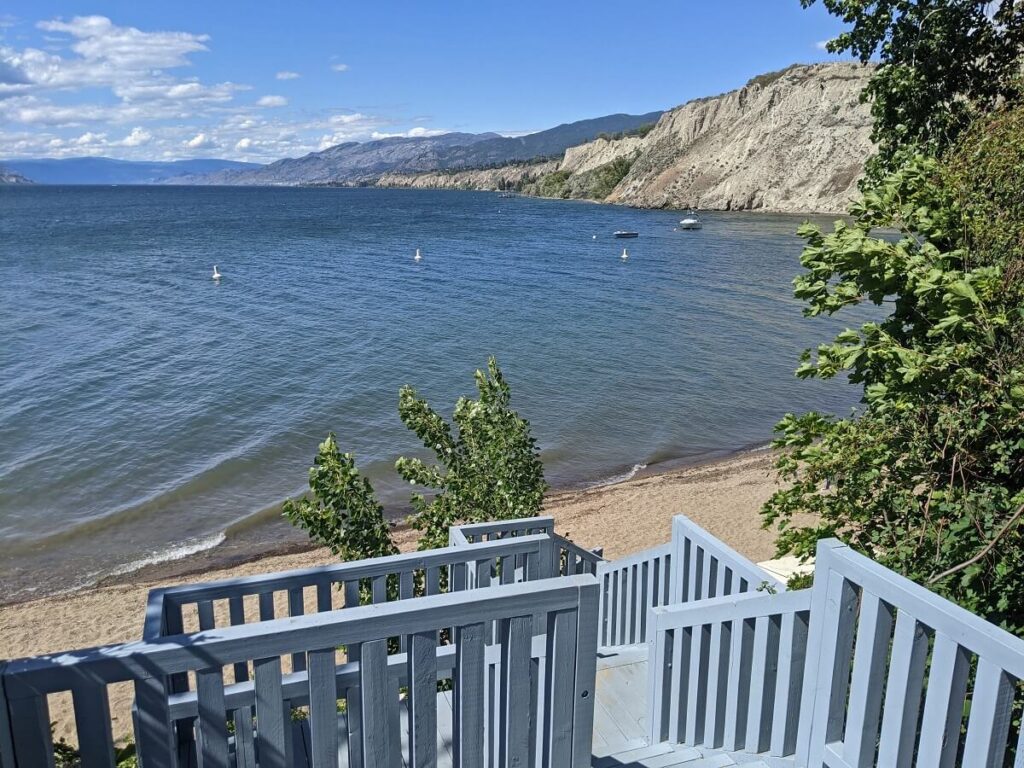 Wooden stairs leading down to sandy beach at Three Mile in Penticton. Huge cliffs rise out of the water on the right hand side. The lake is calm