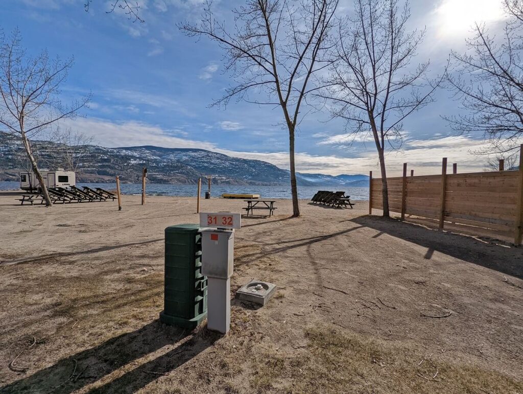 Winter view of Wright's Beach Camp, with white electrical hookup in foreground, area sandy campground area behind, upended picnic tables and bare trees. Skaha Lake and snowy hills are visible in the background