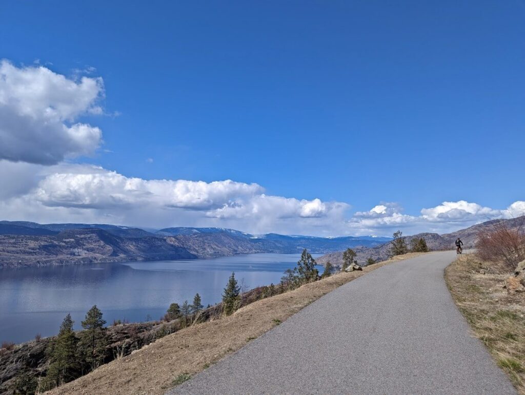 Paved path leading away from camera with cyclist in distance. There is a drop off to the left with Okanagan Lake visible below and forested hills on the other side