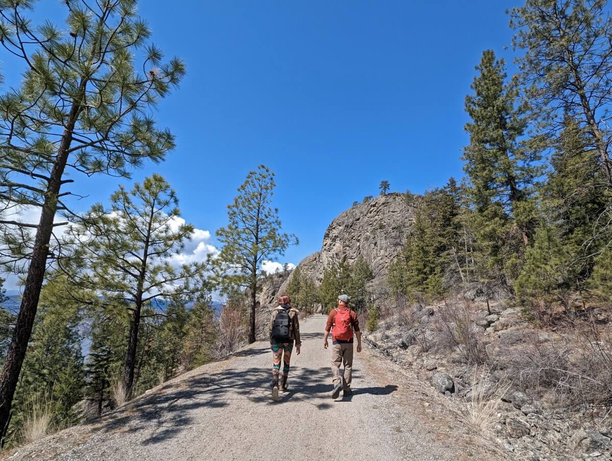 Back view of two hikers on KVR Trail, heading to Little Tunnel. Dry landscape with scattered trees to left and right, with large rock formation in background