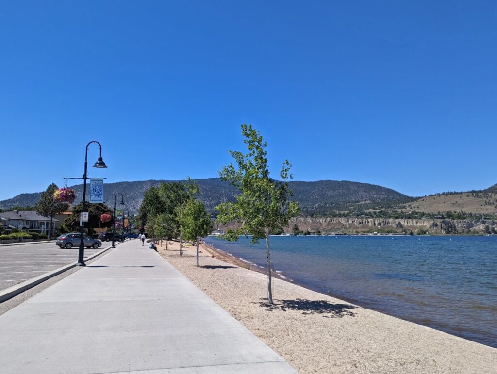 Paved Okanagan Lake promenade stretching away from camera into the distance, with Okanagan Lake on right and parking on left