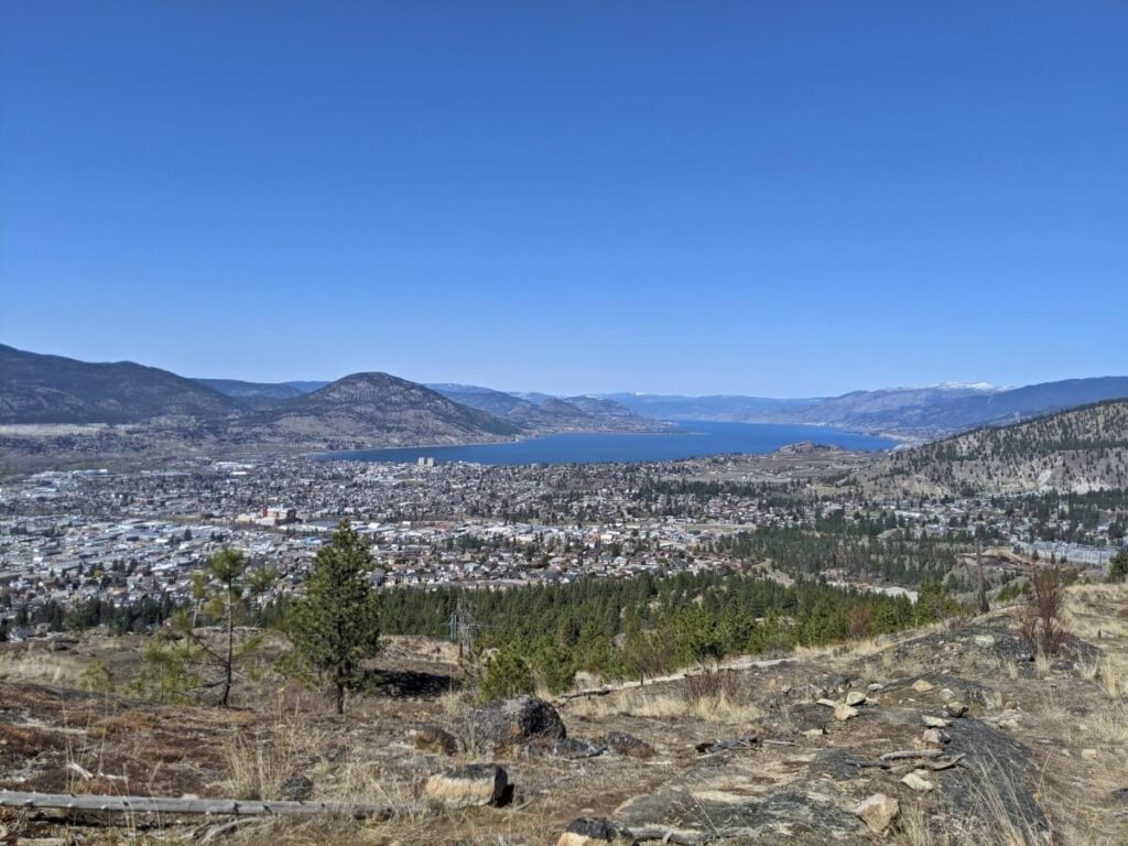 G-Spot hiking trail view looking out to City of Penticton below, with scattered trees in foreground. Okanagan Lake sits on the other side of the town, surrounded by mountains