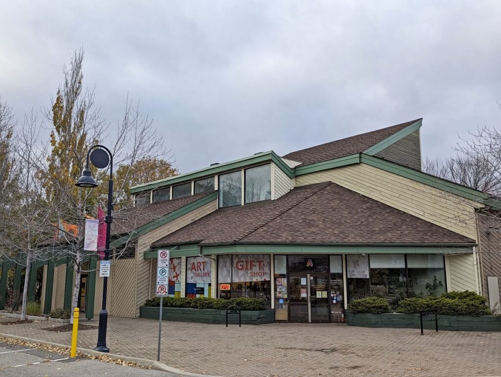 Exterior of Penticton Art Gallery, an unusually shaped two storey building with trees on the left and bushes in front