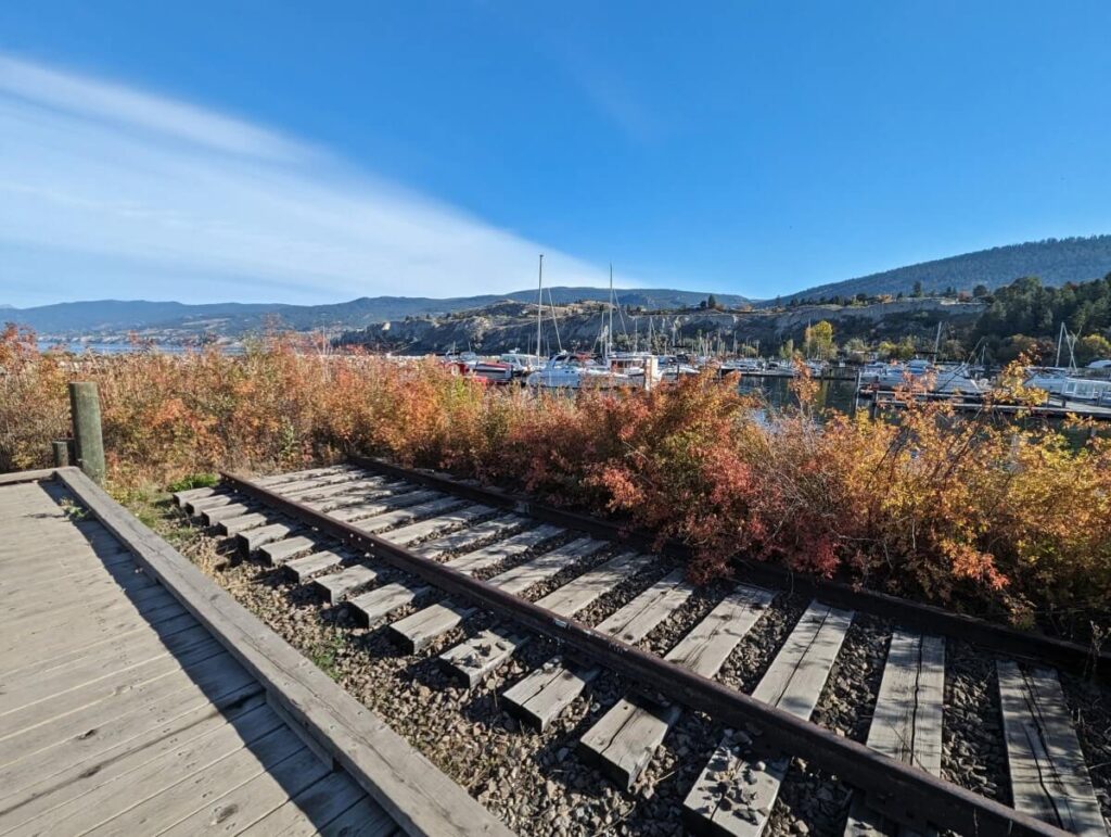 KVR train tracks next to boardwalk near Penticton Marina (visible in background), surrounded by autumnal coloured bushes