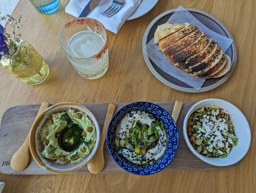 Overhead view of three mezze dips at Elma restaurant with sliced bread above, cockail and vase of flowers on left
