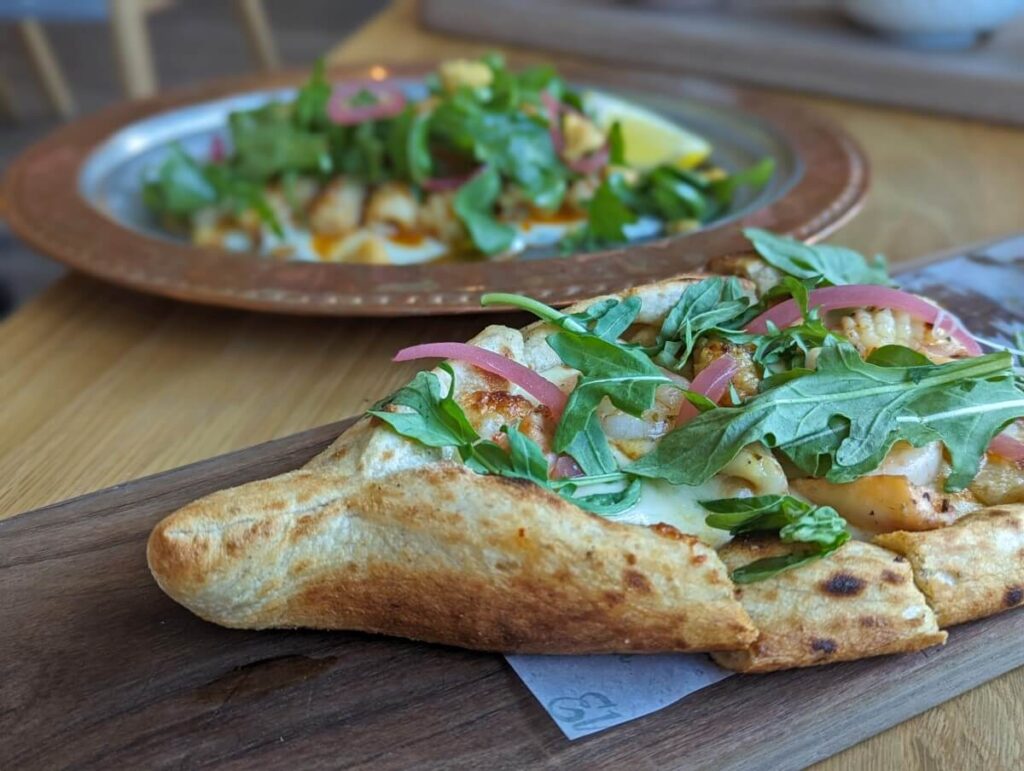 Close up of pide (Turkish flat bread) at Elma, on wooden board, with cheese, rocket (argula) and pickled onions visible. Another dish, also topped with rocket, is visible in the background