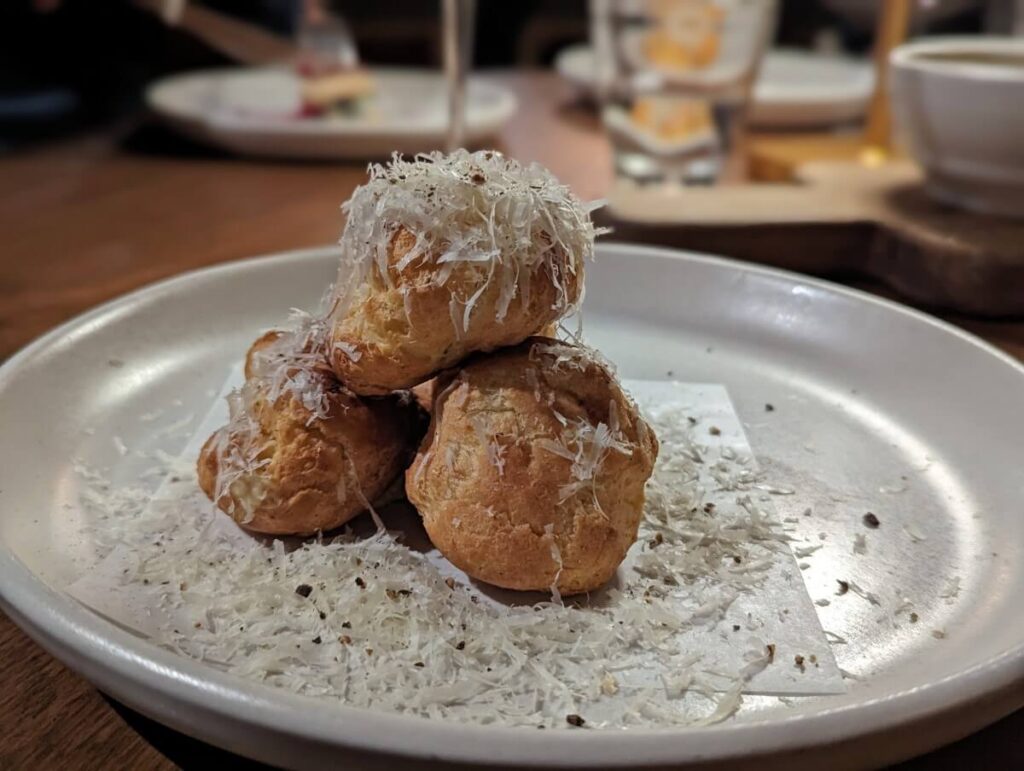 Close up of black pepper gougeres at Eliza, featuring small tower of four pastries on plate, scattered parmesan cheese 