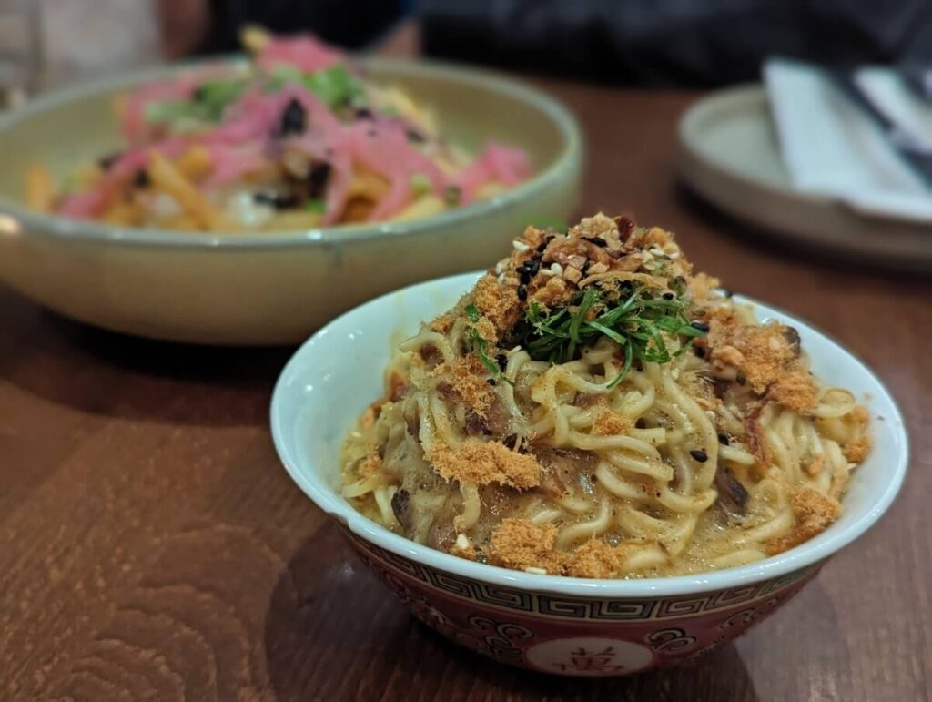 Close up of Kin & Folk 'Carbonara' dish in small round dish noodles and sauce, pork 'floss' and sesame seeds on top. Another dish, topped with pickled onions, is visible in background