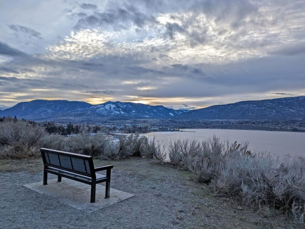 Back view of bench on KVR Trail, looking out to Okanagan lake and the city of Penticton, with snow covered surrounding mountains