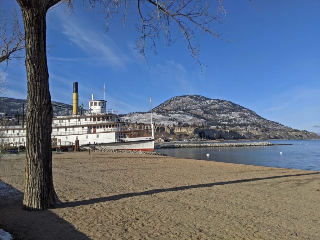Sicamous paddle wheeler on Penticton beach with grey skies and bare tree in foreground. The beach is sandy, even though it is winter. The mountain in the background has snow on it
