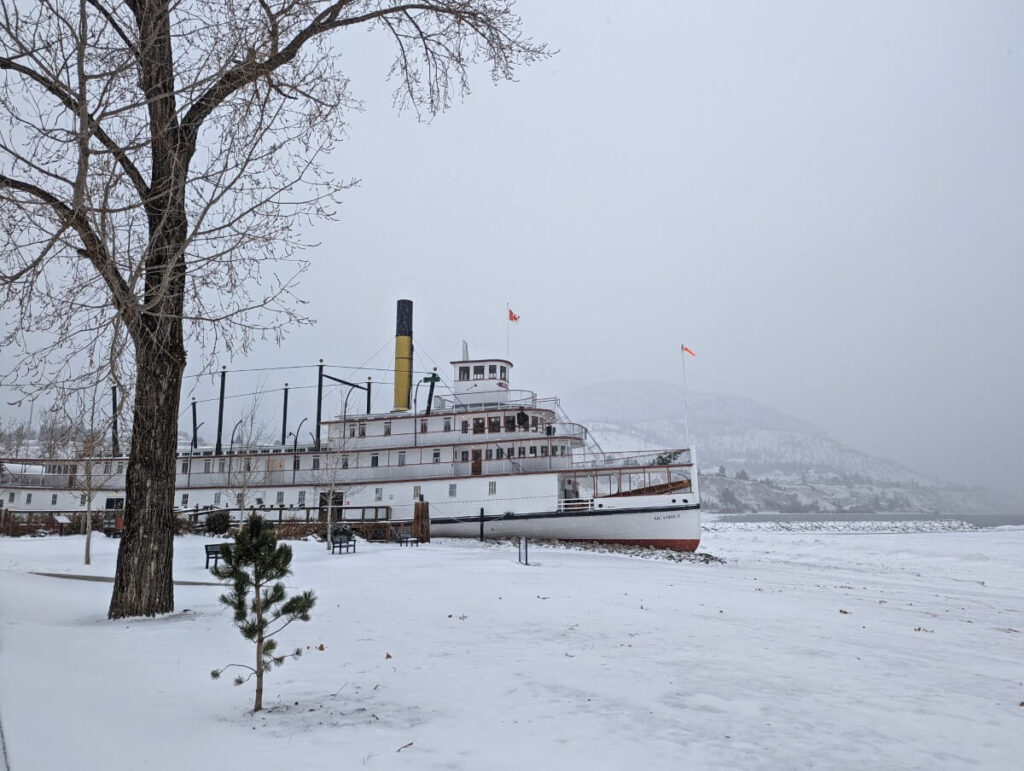 Sicamous paddle wheeler on snowy Penticton beach with grey skies and bare tree in foreground