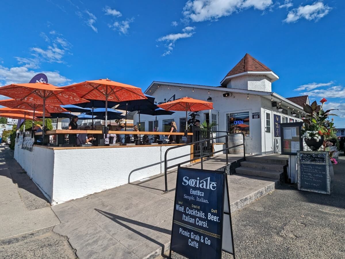 Exterior of Socialē Enoteca Ristorante, single storey white building with large patio (orange umbrellas) on left, signage in front