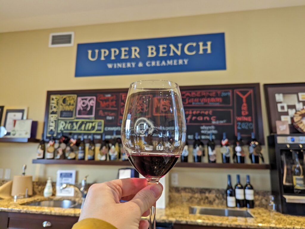 A hand holds up a tasting glass of red wine in front of Upper Bench Winery and Creamery signage