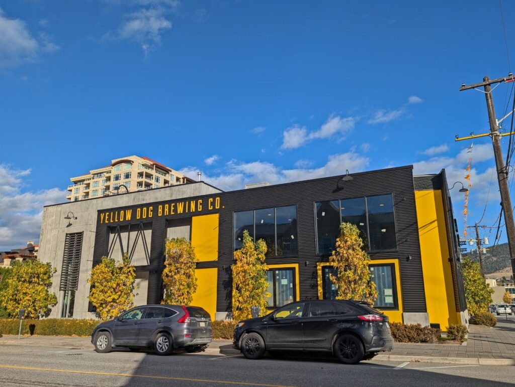 Sidewalk view of Yellow Dog Brewing Co, which is a two storey modern black and yellow building in downtown Penticton