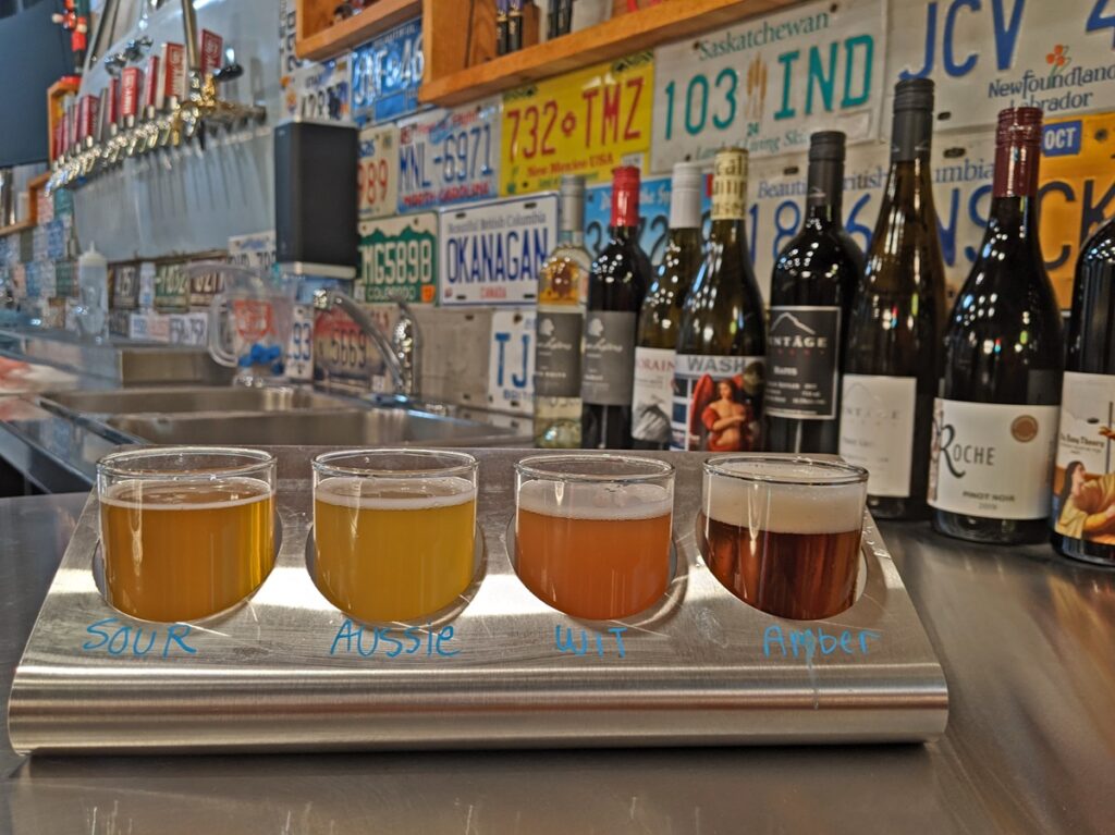 Beer flight with range of beers at Highway 97, set on counter with wine bottles and many licence plates (wall decoration) in background