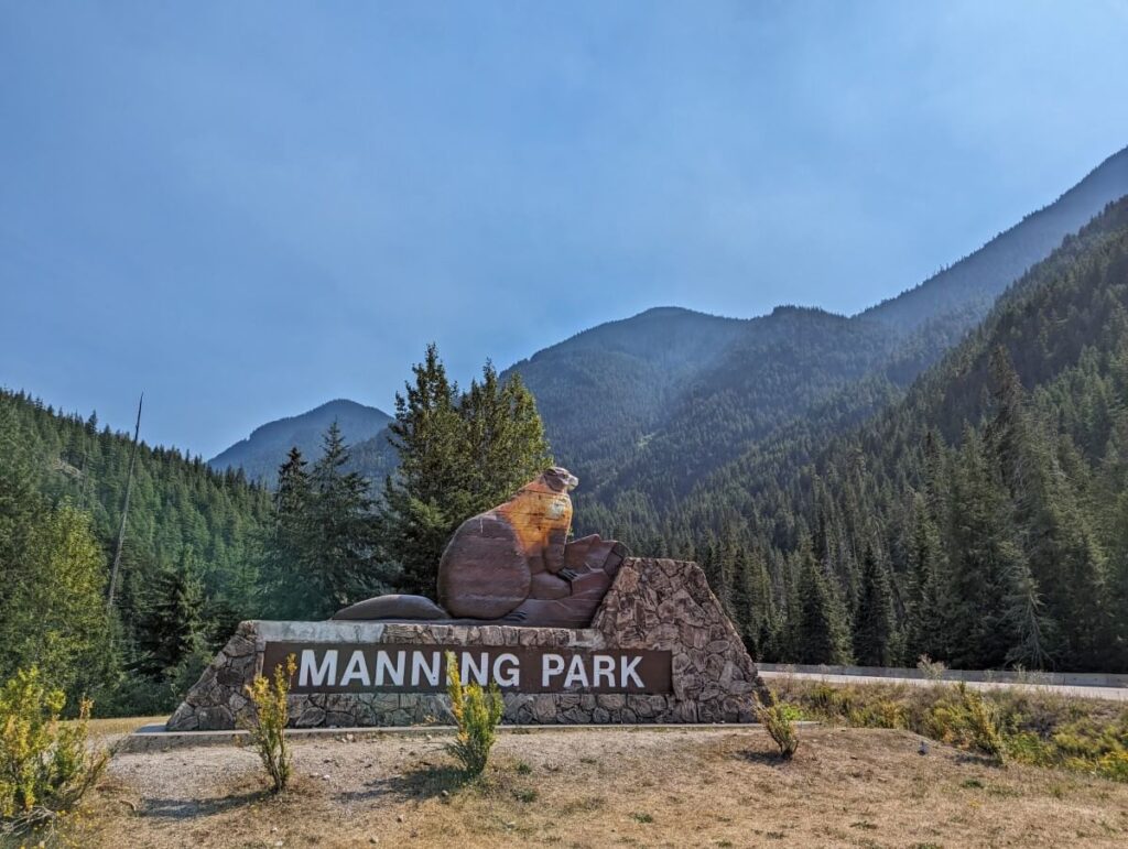 Stone and wooden Manning Park entrance sign with large marmot featured above name. Forested mountains in background