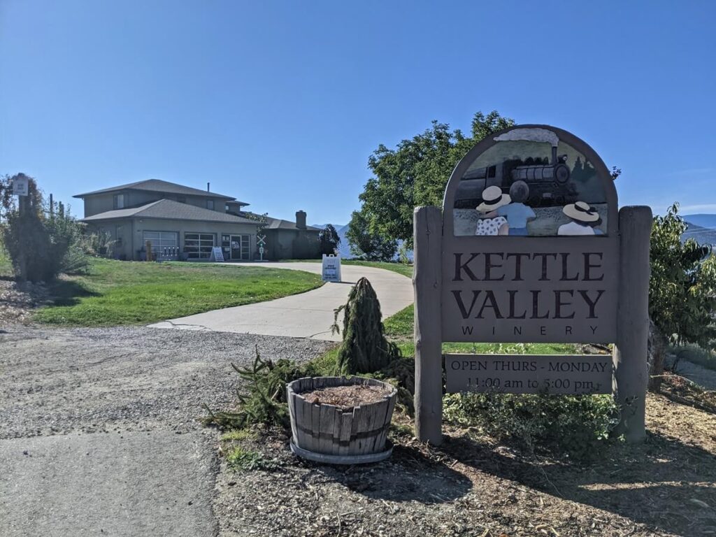 Kettle Valley Winewry sign on right with operating hours, two storey building visible at end of driveway on left 