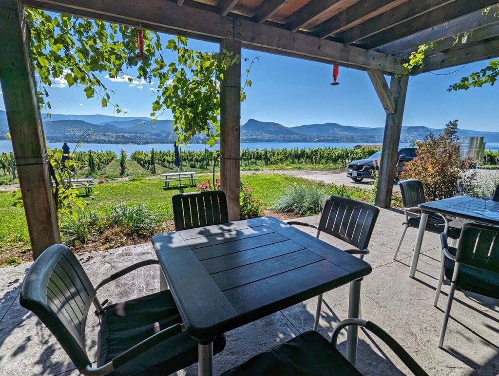 Two sets of table and chairs underneath cover on Deep Roots Winery patio, with lawn area in front and vineyards behind, Okanagan Lake visible in background