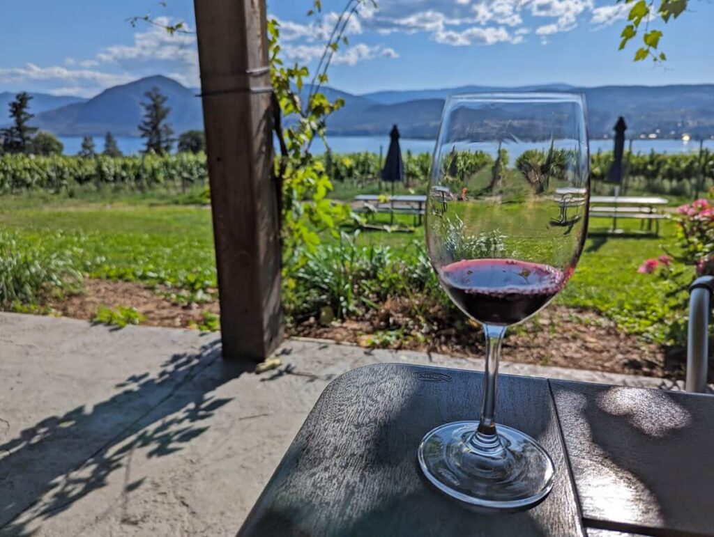 Close up of red wine glass on table in front of lawn and vineyard view, with Okanagan Lake visible in background