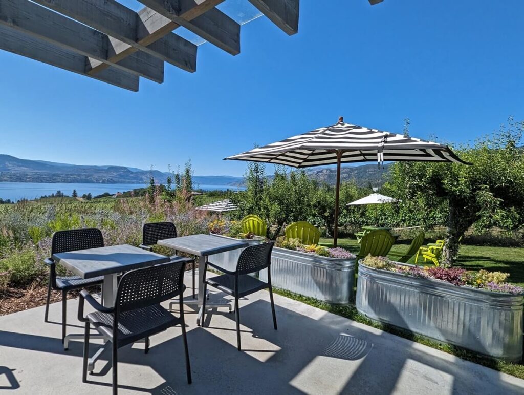Patio seating at Joie Farm, separated from lawn area with planters. Yellow seating is visible on lawn, with umbrellas above. Okanagan Lake can be seen in the background