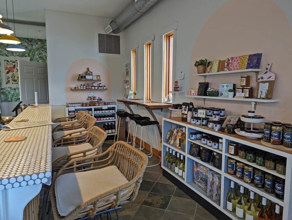 Side bar with bar seating on left, display cases with picnic supplies on right in Poplar Grove Cheese tasting room. White walls, low hanging lights