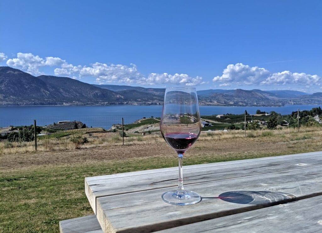 Tasting glass of red wine on wooden picnic table at Origin Winery, with Okanagan Lake views and mountains in background