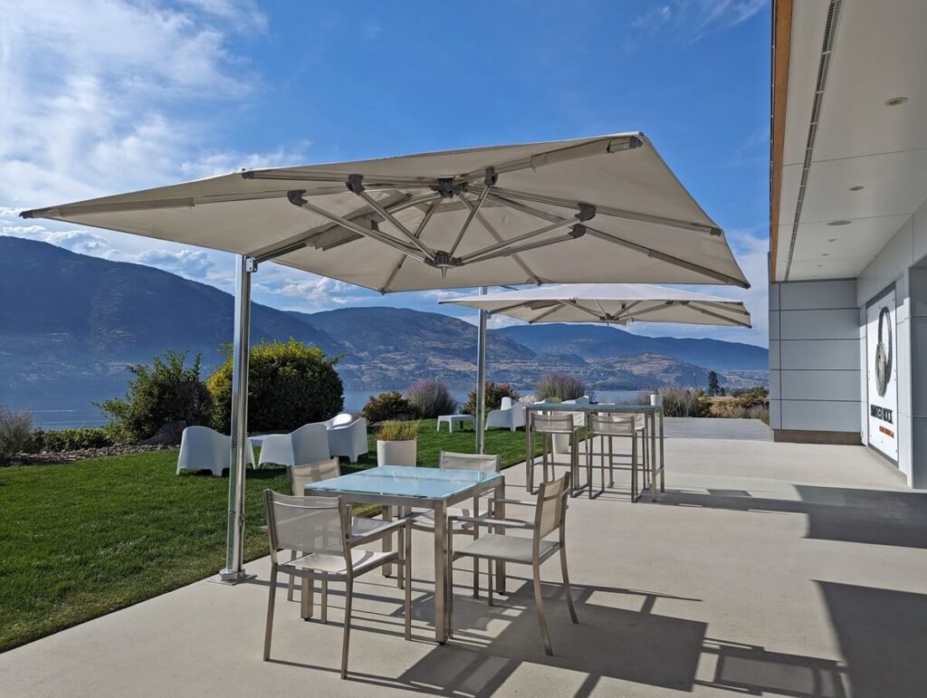 Looking across Painted Rock Winery patio with huge white umbrellas, white coloured seating and tables, lawn on left, modern tasting room on right, lake and forested mountains visible in background