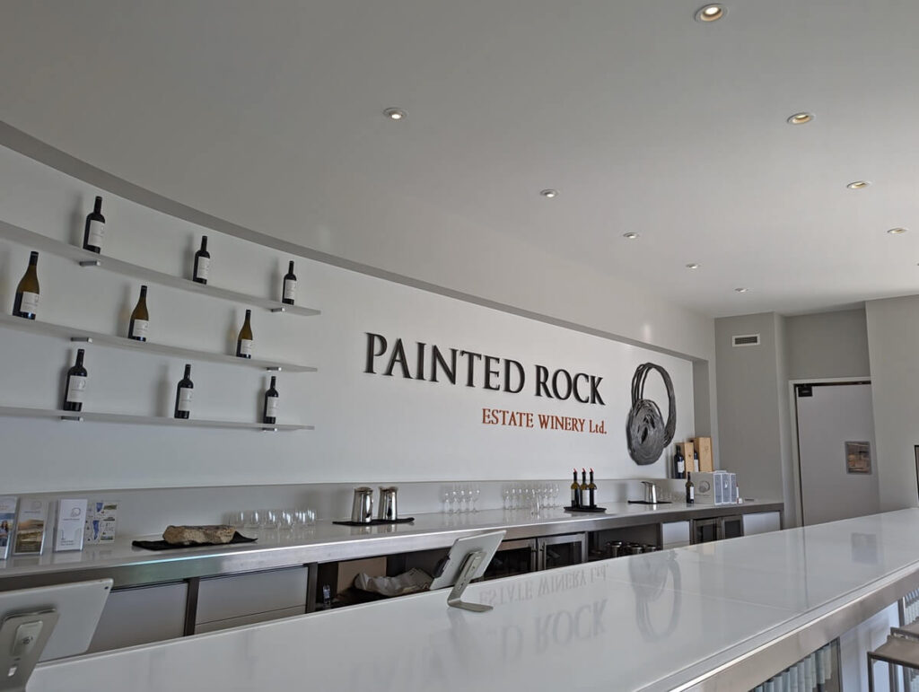 Painted Rock Estate Winery tasting room view across clean white modern bar, with logo on back white wall, with wine bottles on shelves