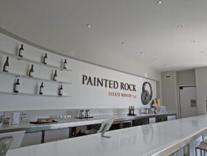 Painted Rock Estate Winery tasting room view across clean white bar, with logo on back white wall, with wine bottles on shelves