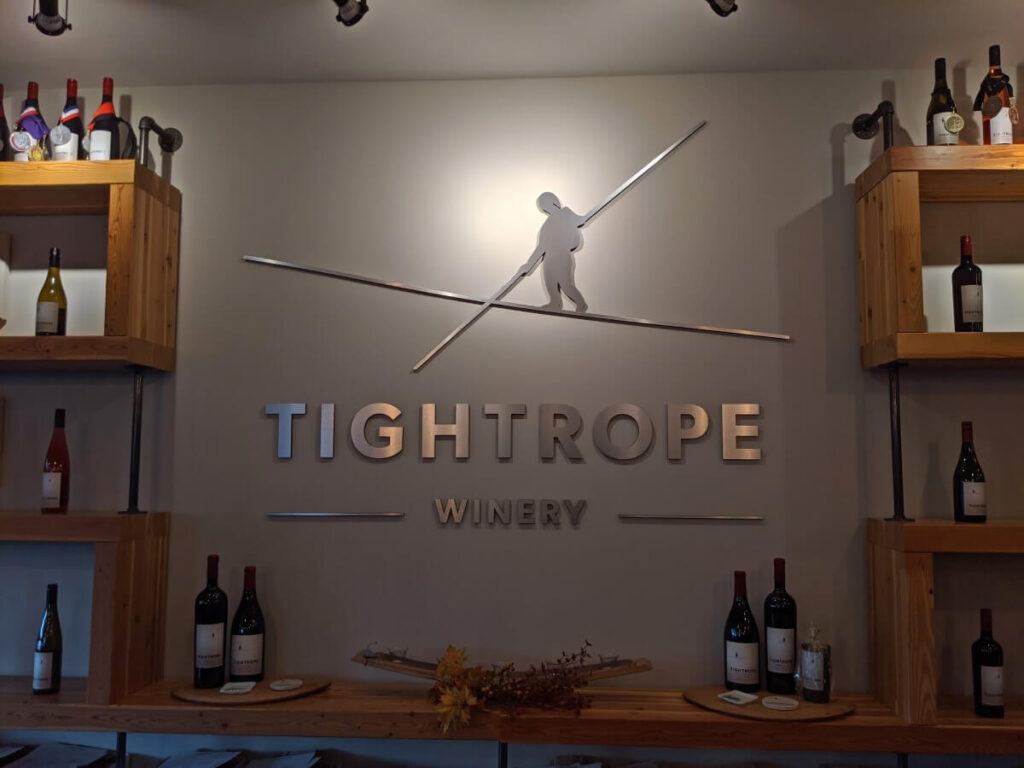Tightrope winery tasting room logo with person balancing on tightrope. Wooden shelves surround the large sign with wine bottles scattered