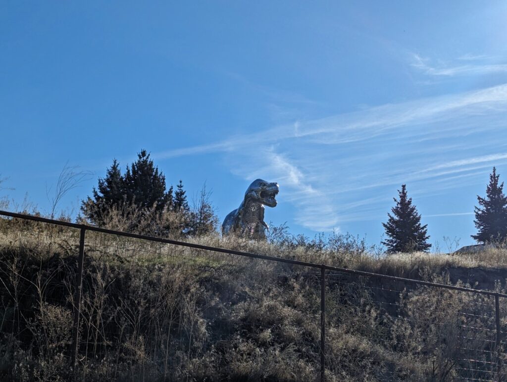 Looking up at metal Alice the T-Rex sculpture over metal fence from the KVR Trail in Penticton