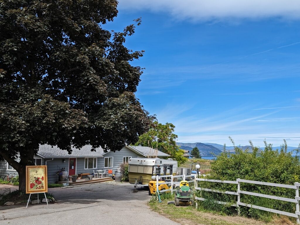 Looking down to driveway slope with Fox and Archer sign on left, single story house in middle, trailer on right and tractor next to fence on far right. Okanagan Lake is visible in the background