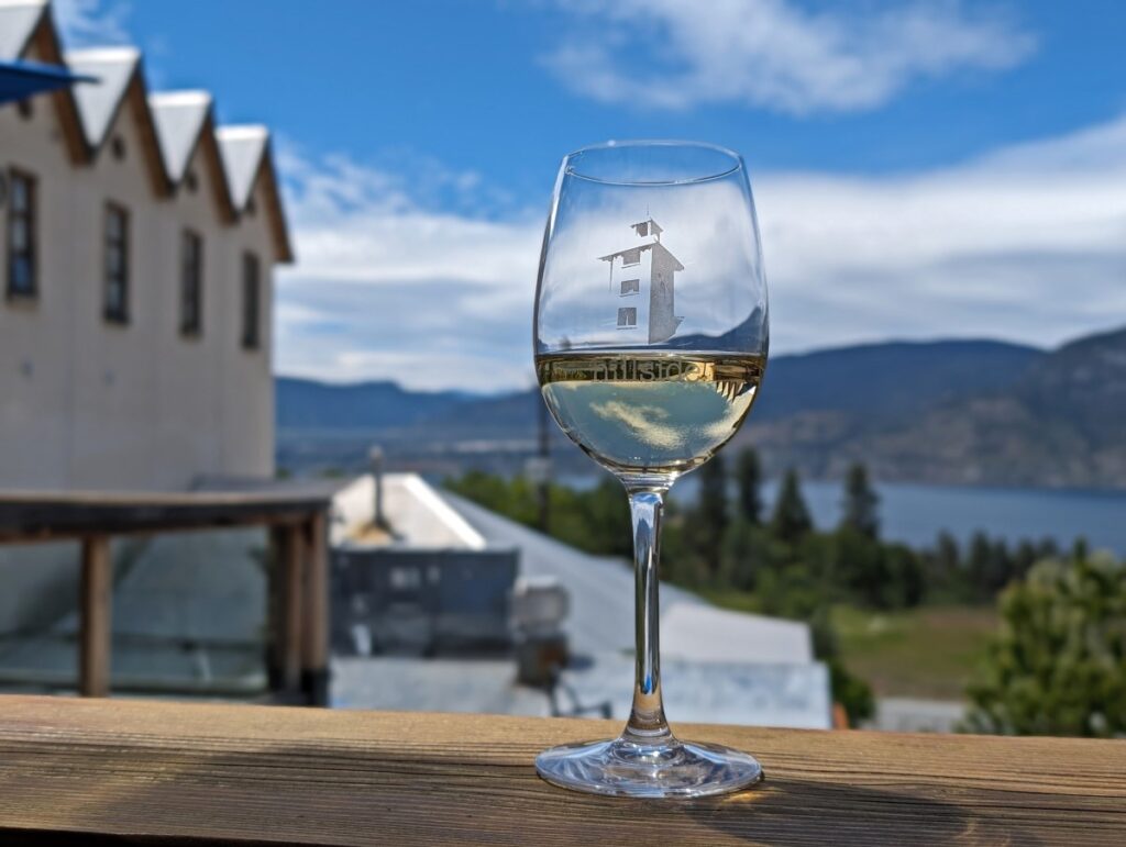 Close up of wine glass containing white wine on wooden surface in front of Hillside Winery building view, featuring Okanagan lake in background