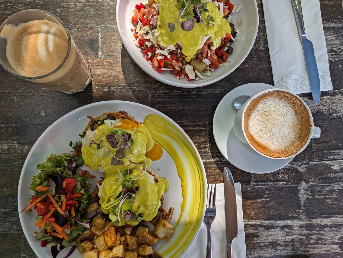 Overhead shot of colourful vegan Eggs Benny and Mexican Fiesta dishes at Stillfood Bistro with two coffees on either side with knives and forks